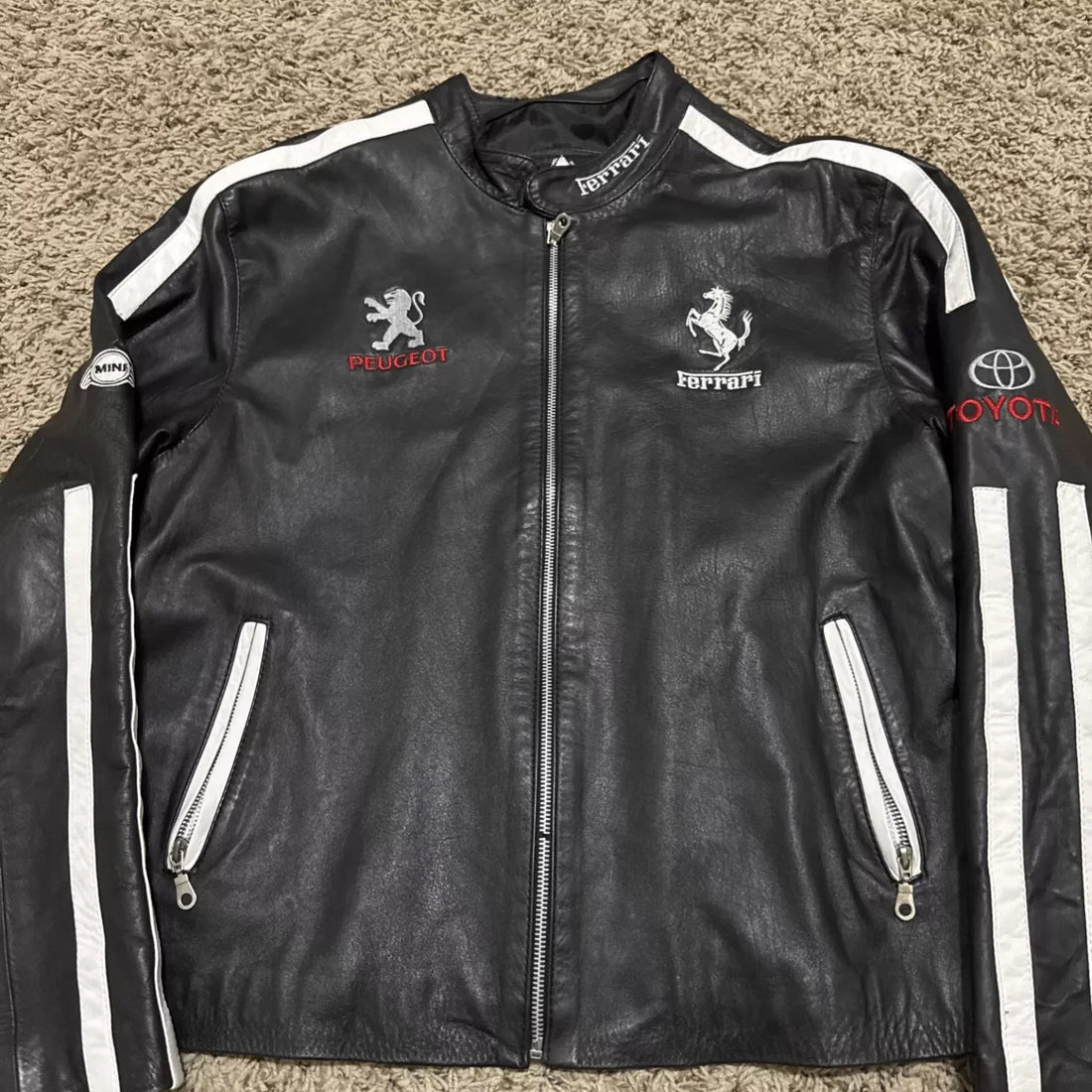 Full front view of a black leather multi-brand motorsport jacket with white stripes, featuring Peugeot, Ferrari, Toyota, and MINI logos.