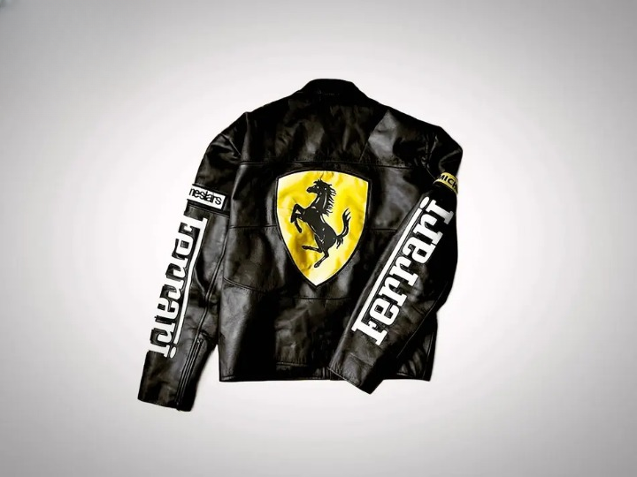 A black leather Ferrari Racing jacket with large white Ferrari text on the chest and sleeves, featuring small Bell, Vodafone, and Michelin sponsor patches.